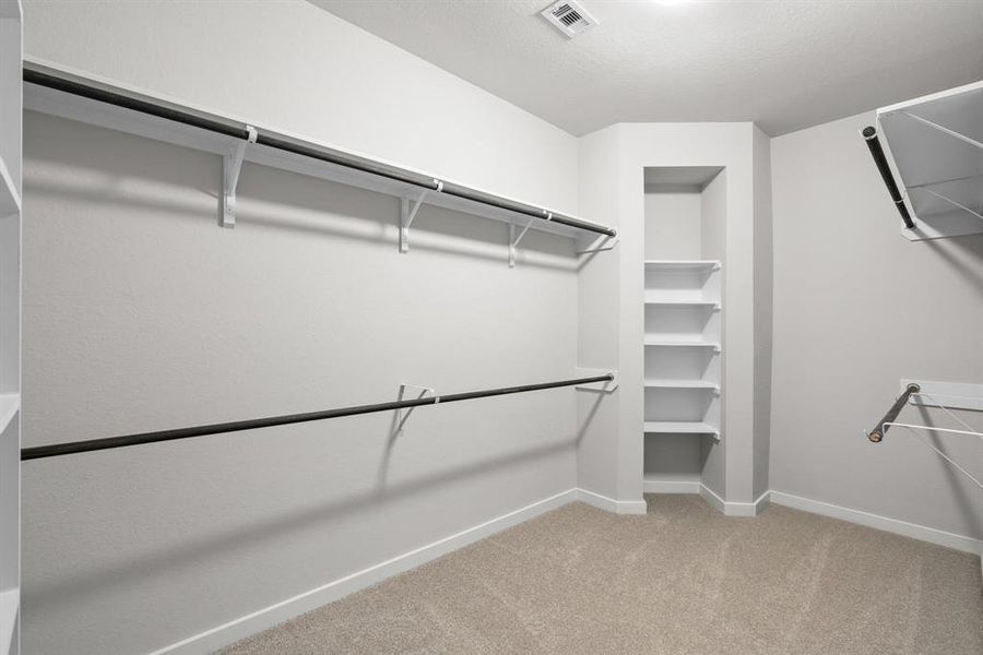 Experience luxury in this spacious walk-in closet with high ceilings and plush carpet. Warm paint tones, built-in shelving, and dark finishes create a contemporary and functional retreat. Sample photo of completed home with similar floor plan. As-built interior colors and selections may vary.