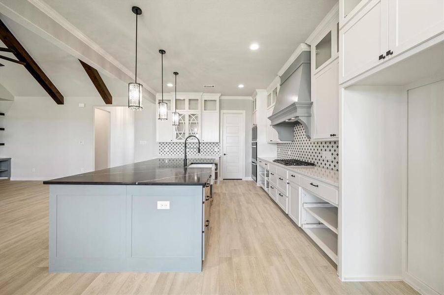 Kitchen with custom exhaust hood, tasteful backsplash, white cabinets, light wood-type flooring, and an island with sink