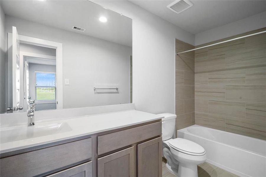 Secondary bath features tile flooring, bath/shower combo with tile surround, stained wood cabinets, beautiful light countertops, mirror, sleek fixtures and modern finishes.