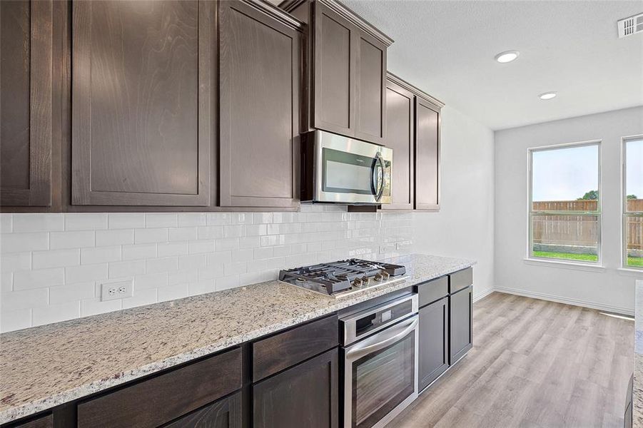 Kitchen with light hardwood / wood-style flooring, dark brown cabinets, light stone counters, stainless steel appliances, and decorative backsplash