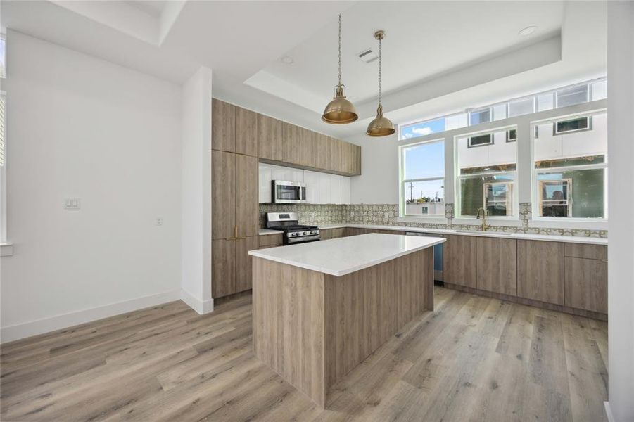 Modern and bright kitchen with light cabinetry, a central island, stainless steel appliances, and sleek pendant lighting. The room features large windows allowing for natural light, and has upgraded flooring throughout.