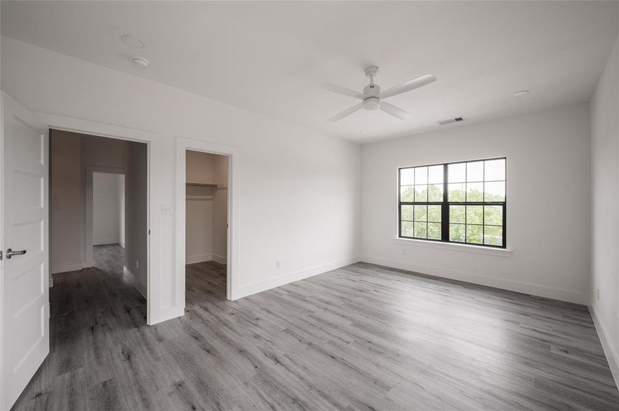 *Representative Photo.Spacious Primary Bedroom featuring modern grey plank flooring, fresh white walls, a large window, and a ceiling fan. The room includes a spacious walk in closet