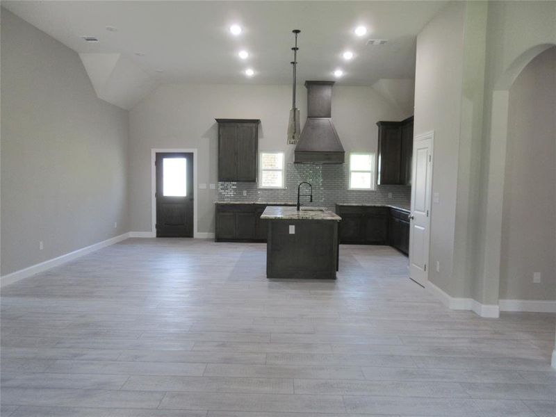 Kitchen with custom range hood, light stone counters, a kitchen island with sink, decorative backsplash, and vaulted ceiling