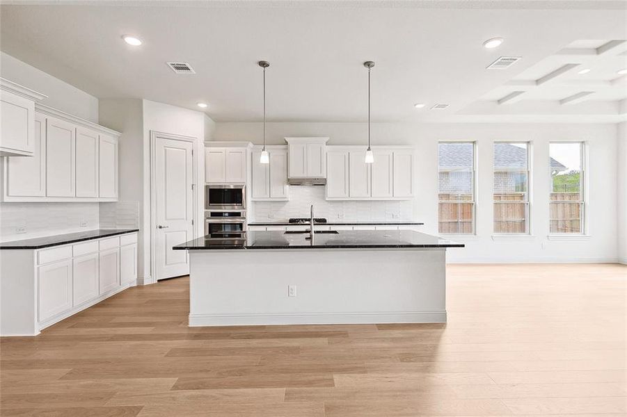 Kitchen with light hardwood / wood-style floors, stainless steel microwave, a kitchen island with sink, decorative light fixtures, and tasteful backsplash