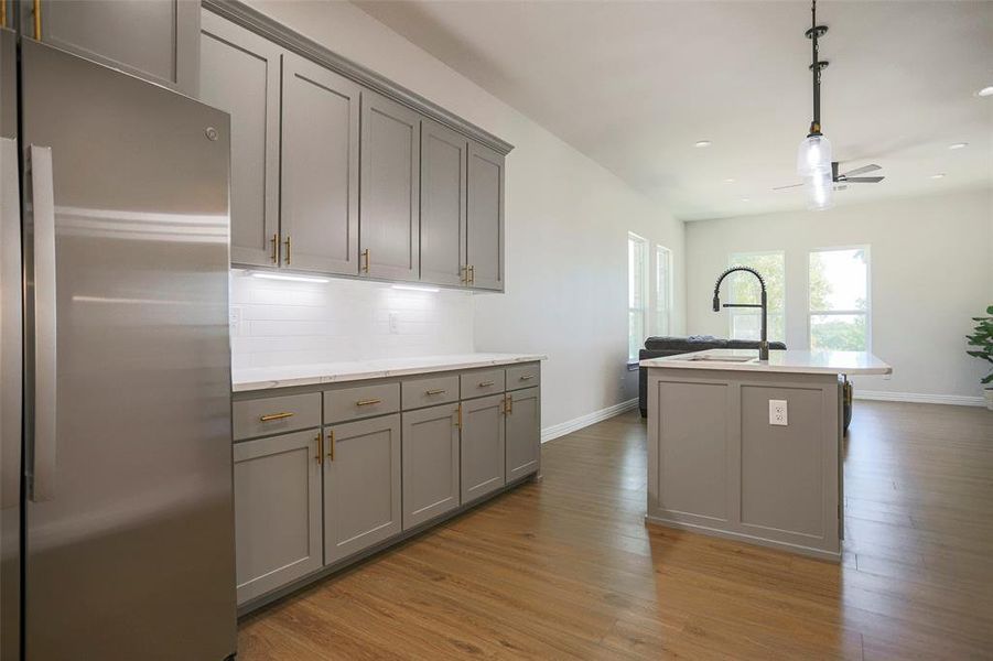 Kitchen featuring light wood-type flooring, stainless steel refrigerator, a center island with sink, and gray cabinets. Soft close cabinets and drawers throughout.