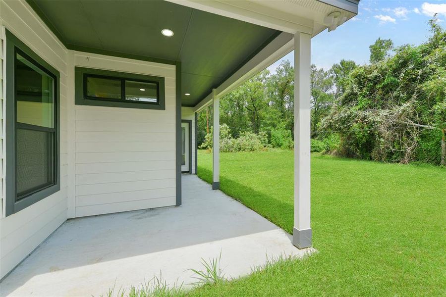 The photo shows the exterior side of a modern home featuring a covered patio area with recessed lighting and a view of a well-maintained lawn.