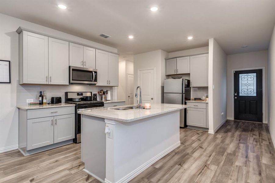 Kitchen with light stone counters, tasteful backsplash, a kitchen island with sink, stainless steel appliances, and light hardwood / wood-style floors