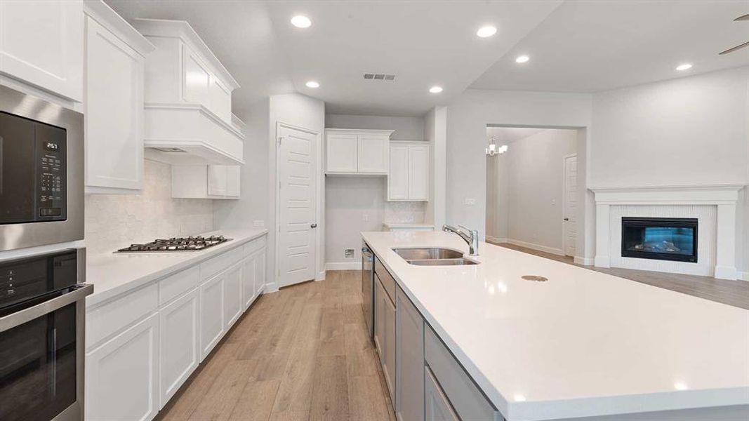 Kitchen with light wood-type flooring, backsplash, appliances with stainless steel finishes, a fireplace, and a center island with sink