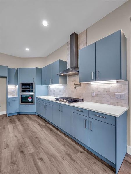 Kitchen with wall chimney range hood, light hardwood / wood-style flooring, blue cabinets, and backsplash