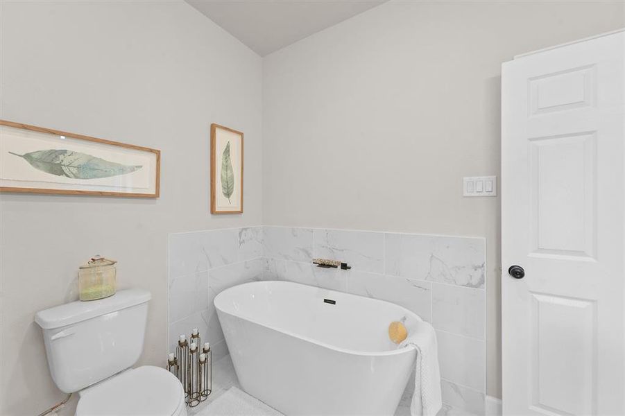 This is a modern bathroom featuring a freestanding bathtub with a marble wall backdrop, a white toilet, and tasteful botanical artwork on the wall. The space is accented with simple, elegant decor and neutral colors.