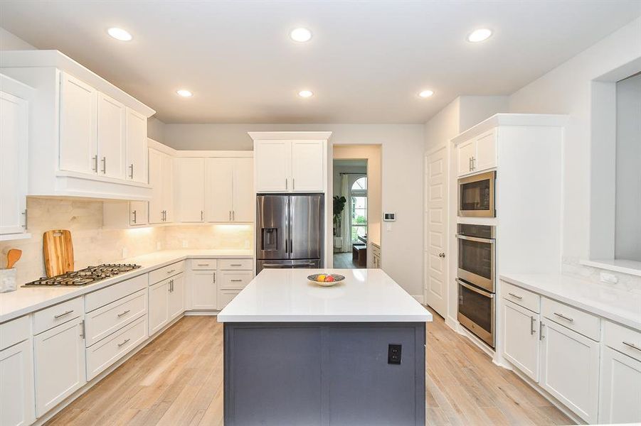 Modern, open-plan kitchen with white cabinetry and a contrasting blue island, featuring stainless steel appliances, recessed lighting, and tile floors. The kitchen opens up to a dining area with ample natural light as well as a large living area.