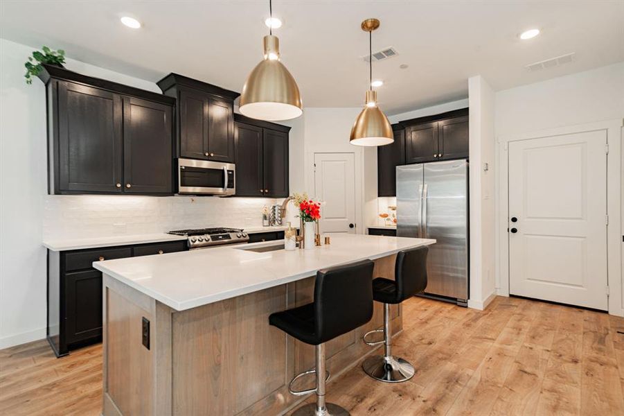 Kitchen with stainless steel appliances, hanging light fixtures, light hardwood / wood-style flooring, backsplash, and a kitchen island with sink