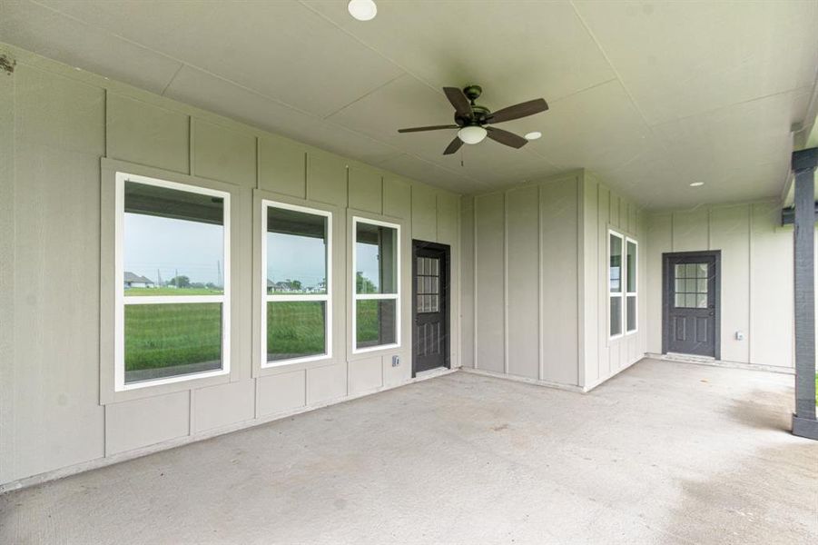 This is a spacious, covered patio area featuring multiple windows providing ample natural light, a ceiling fan for comfort, and two exterior doors for easy access. It overlooks a green, open field, offering a serene outdoor view.