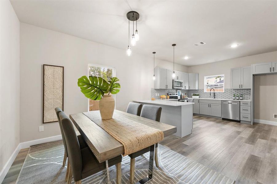 This photo showcases a modern, open-concept dining and kitchen area with sleek finishes. The dining space has a rustic wooden table, while the kitchen features gray cabinetry, stainless steel appliances, and a subway tile backsplash. Natural light and neutral colors create a welcoming atmosphere.