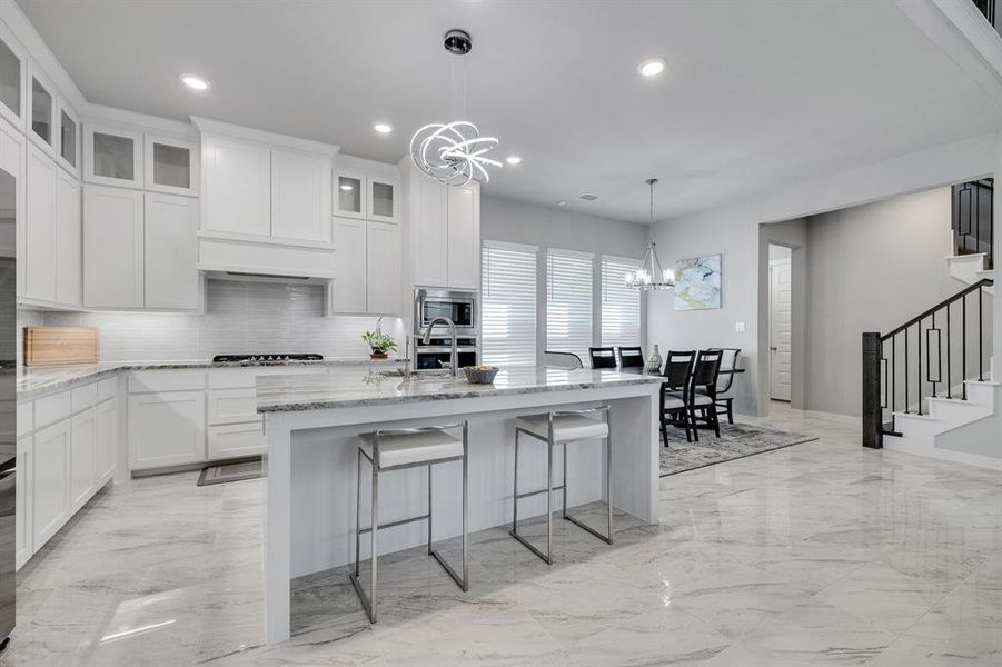 Kitchen with an inviting chandelier, stainless steel microwave, light stone countertops, backsplash, and a kitchen island with sink