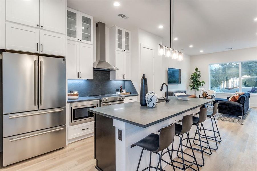 Kitchen with white cabinets, a breakfast bar, wall chimney exhaust hood, stainless steel appliances, and a kitchen island with sink