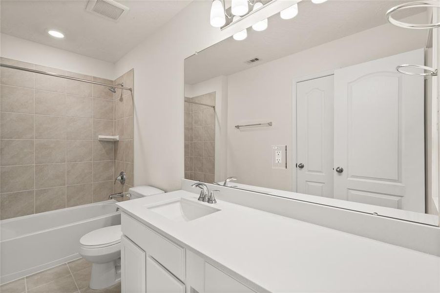 Secondary bath features tile flooring, bath/shower combo with tile surround, white stained wood cabinets, beautiful light countertops, mirror, dark, sleek fixtures and modern finishes.