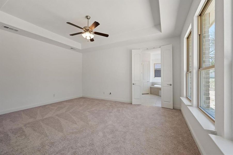 Unfurnished bedroom with a raised ceiling, ensuite bathroom, light colored carpet, and ceiling fan