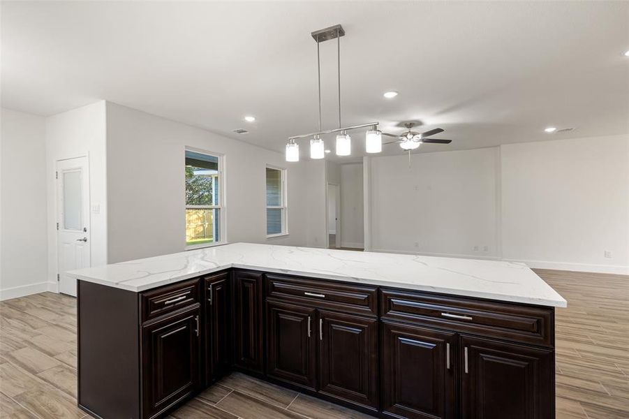 Kitchen with light wood-type flooring, ceiling fan, dark brown cabinets, light stone countertops, and hanging light fixtures