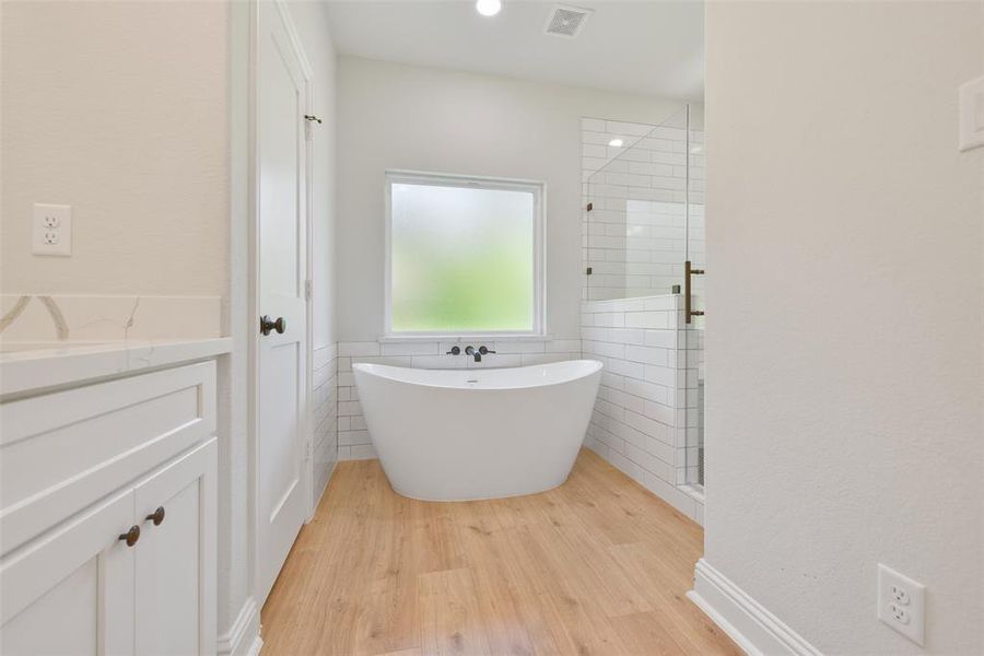 This photo showcases a modern bathroom with a large frosted window providing natural light. It features a stylish freestanding bathtub and a separate glass-enclosed shower, complemented by white subway tiles and a wood-look floor. The vanity double sinks and ample storage with white cabinetry and a quartz countertop.
