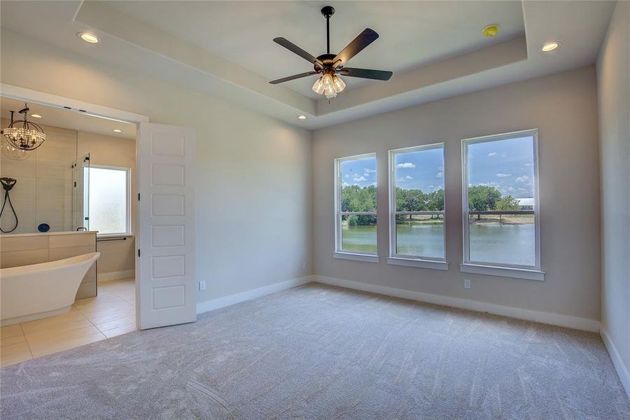 Unfurnished bedroom with light carpet, ceiling fan with notable chandelier, a water view, and a raised ceiling