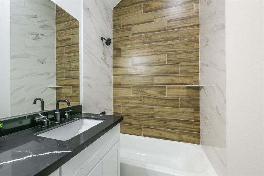 This is a modern bathroom featuring a combination of marble-look and wood-look tile surrounding a built-in bathtub and shower, complete with sleek black fixtures. A white vanity with a dark countertop complements the clean, contemporary design.