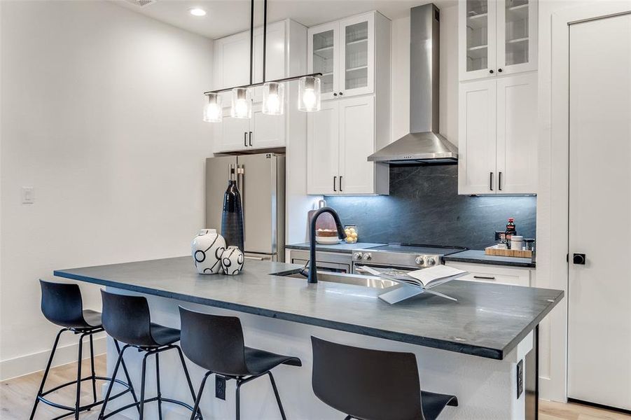 Kitchen with white cabinets, an island with sink, hanging light fixtures, wall chimney range hood, and appliances with stainless steel finishes