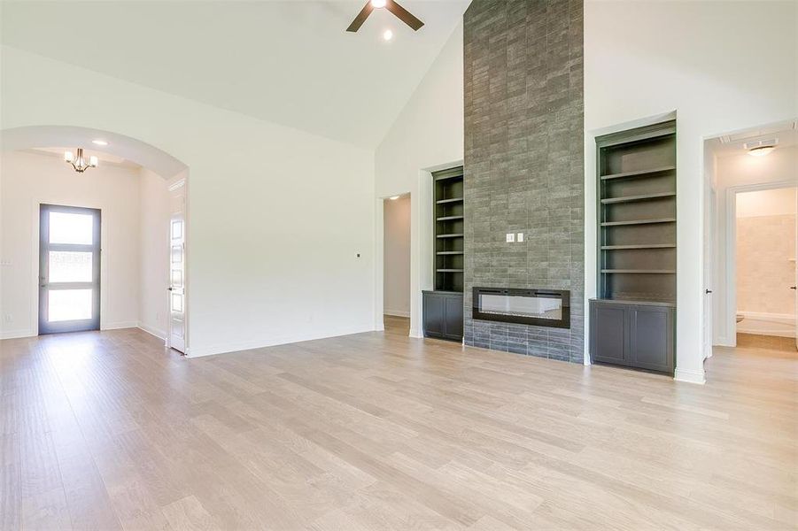 Unfurnished living room with light wood-type flooring, ceiling fan with notable chandelier, built in features, a large fireplace, and high vaulted ceiling