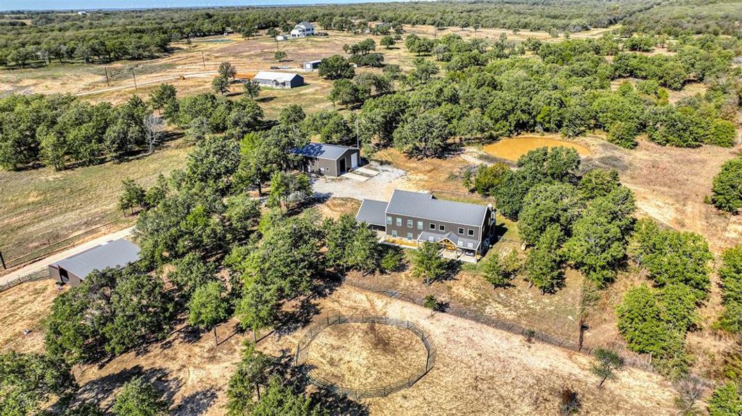 The aerial view captures the expansive barndominium, shop, barn, and serene pond, all nestled among mature trees that create a picturesque and tranquil setting.