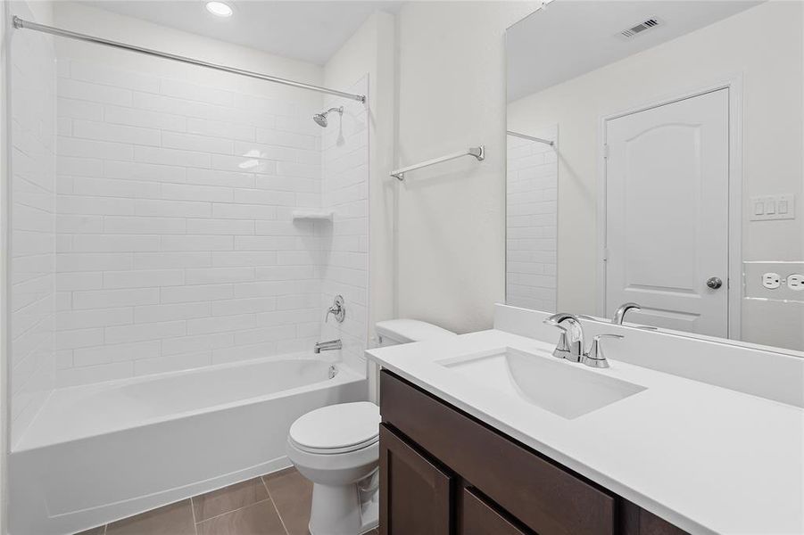 Secondary bath features tile flooring, bath/shower combo with tile surround, stained wood cabinets, beautiful light countertops, mirror, dark, sleek fixtures and modern finishes.