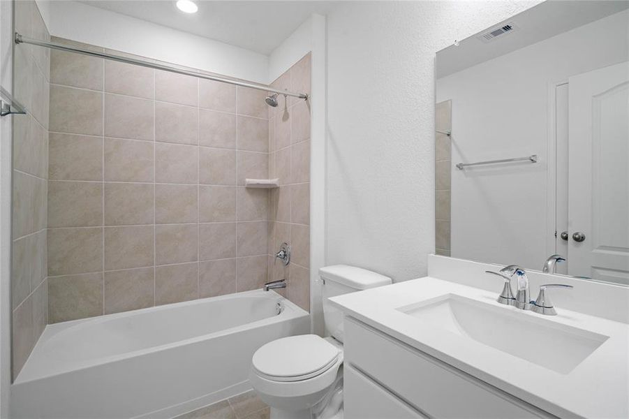Secondary bath features tile flooring, bath/shower combo with tile surround, stained wood cabinets, beautiful light countertops, mirror, dark, sleek fixtures and modern finishes.