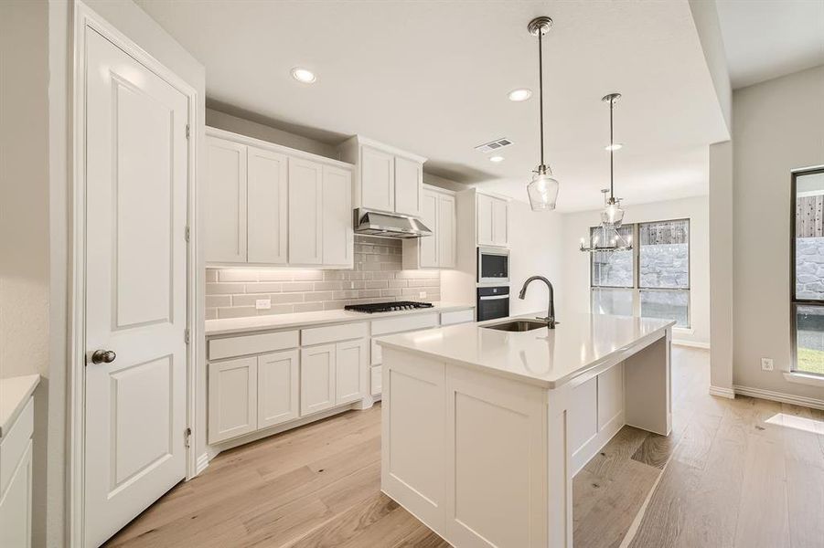 Kitchen featuring light wood-type flooring, an island with sink, stainless steel appliances, decorative light fixtures, and white cabinets