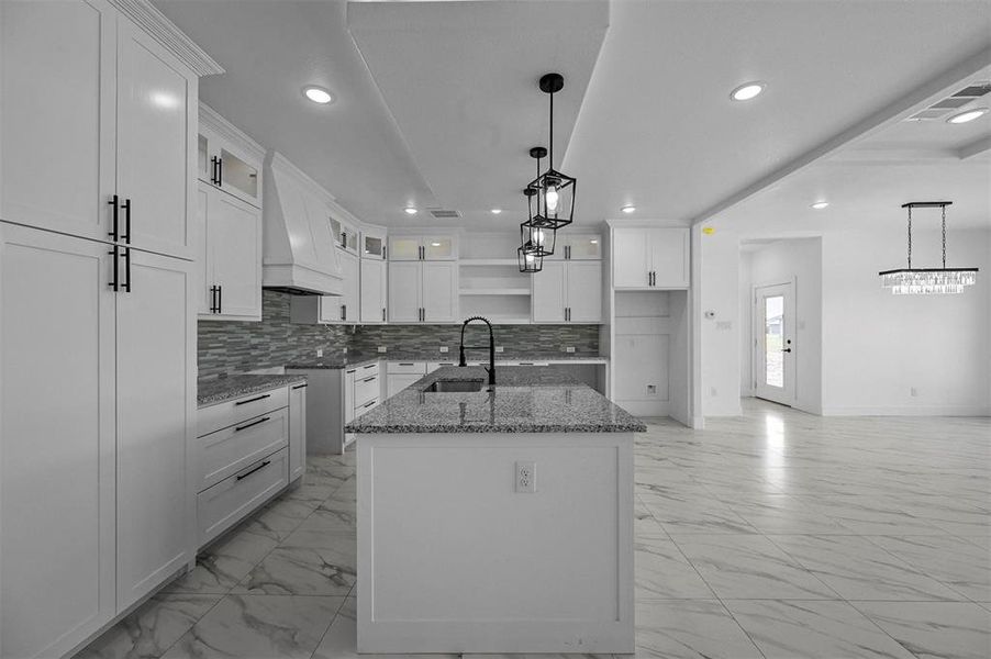 Kitchen featuring white cabinets, premium range hood, an island with sink, pendant lighting, and sink