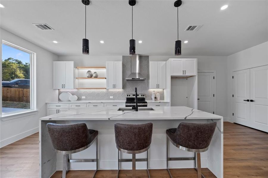 Kitchen featuring a center island with sink, wall chimney range hood, hanging light fixtures, and white cabinets