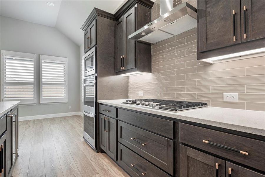 Kitchen with tasteful backsplash, dark brown cabinets, light wood-type flooring, lofted ceiling, and wall chimney exhaust hood