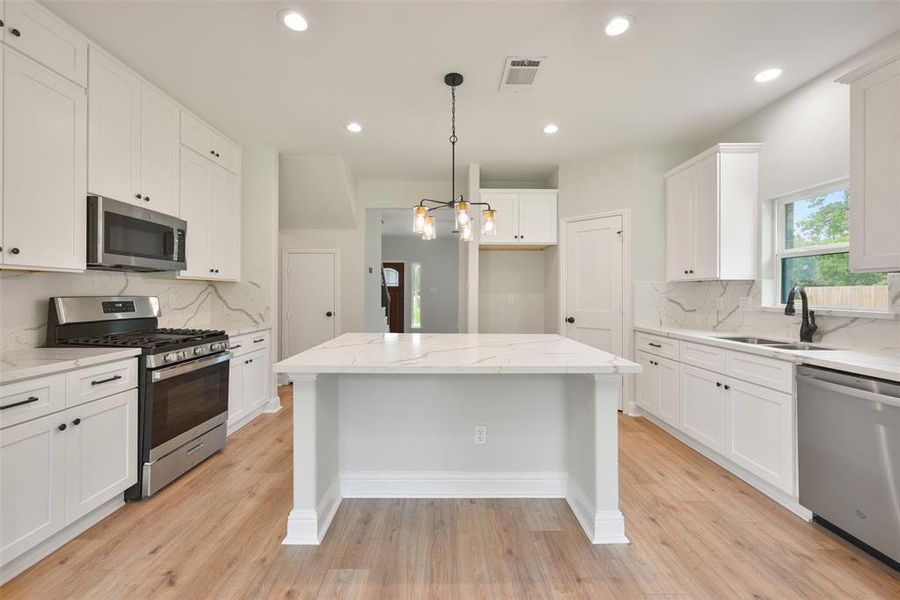 Bright and modern kitchen featuring white cabinetry, premium quartz countertops, stainless steel appliances, and a central island. The space is complemented by elegant light fixtures and natural light from the window.