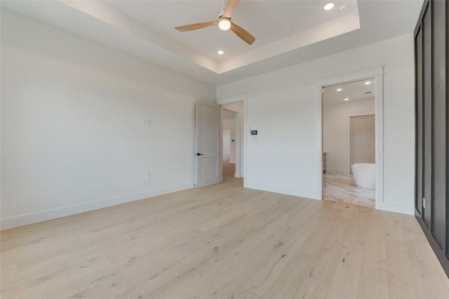 Unfurnished bedroom with light hardwood / wood-style flooring, ensuite bathroom, ceiling fan, and a tray ceiling