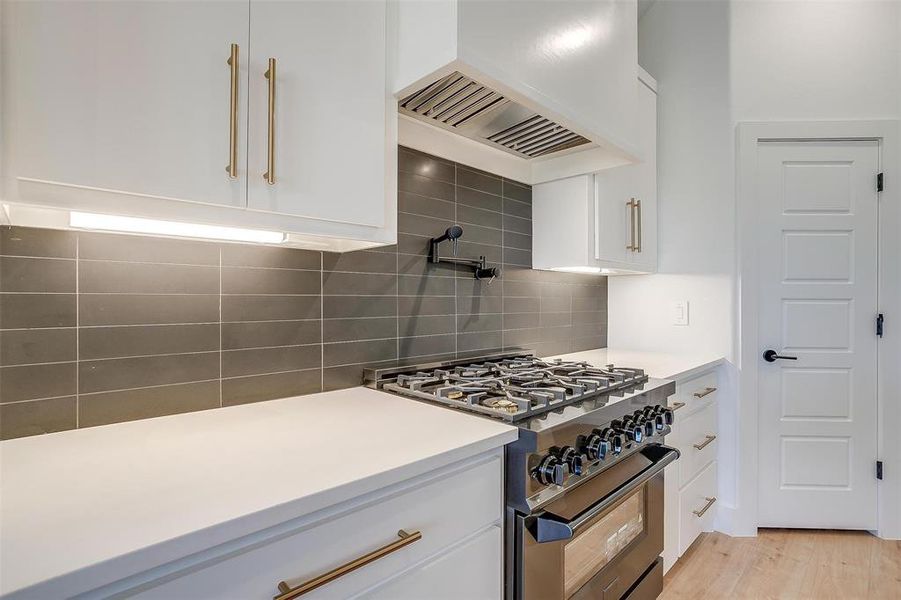 Kitchen featuring light wood-type flooring, stainless steel gas range oven, custom range hood, backsplash, and white cabinetry