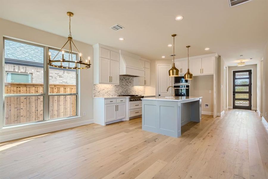 Kitchen featuring a kitchen island with sink, white cabinets, hanging light fixtures, custom range hood, and light hardwood / wood-style flooring