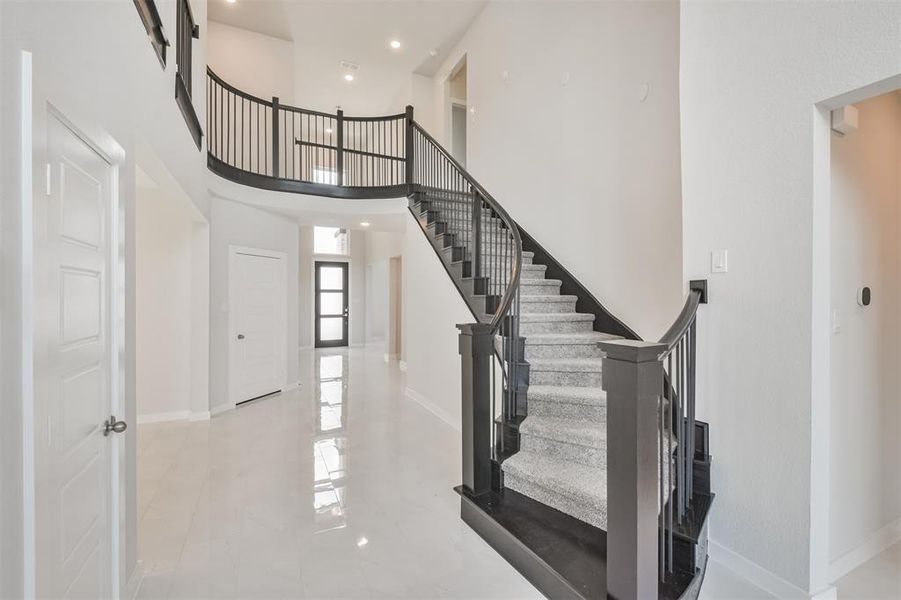 The photo shows a modern entryway with a sweeping staircase featuring dark wood and metal railings, leading to an upper level with a curved balcony. The space is bright with white walls and high ceilings.