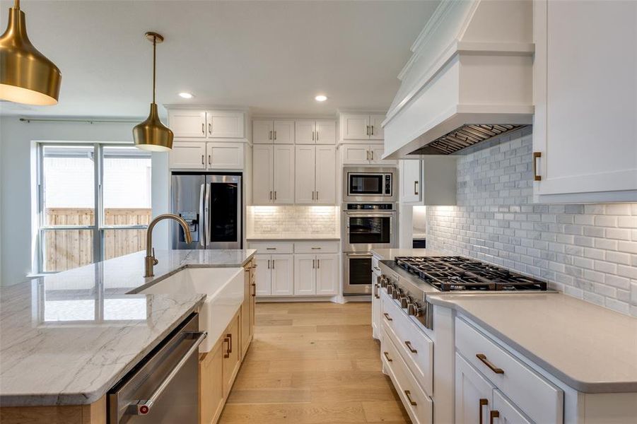 Kitchen featuring an island with sink, custom exhaust hood, appliances with stainless steel finishes, decorative backsplash, and decorative light fixtures