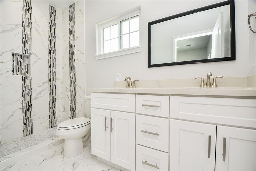 This Primary bathroom features a modern design with a marble-themed tile floor and accent wall, a white vanity with ample storage, double sinks with elegant fixtures, and a large framed mirror. There's also a window providing natural light and a toilet adjacent to a glass-enclosed shower.