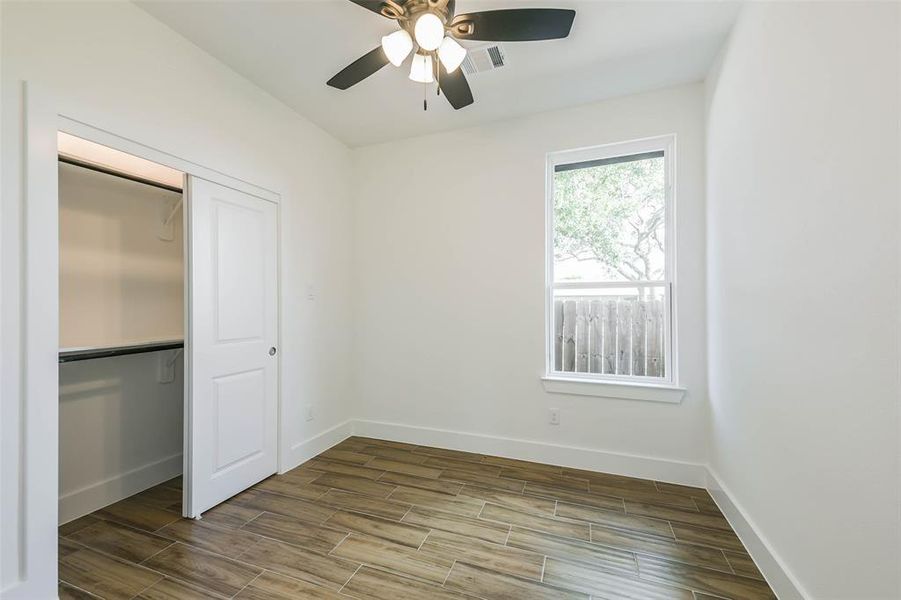 This is a bright, empty room with fresh, neutral walls and modern wood-look flooring. It features a ceiling fan, a window with a view of trees, and a built-in closet space.