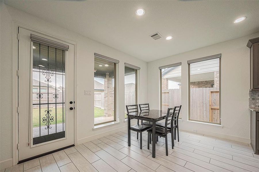 This is a bright, modern dining space featuring large windows, a glass-paneled door, and recessed lighting. It includes a dining set for four and tiled flooring, with a view of a fenced backyard.