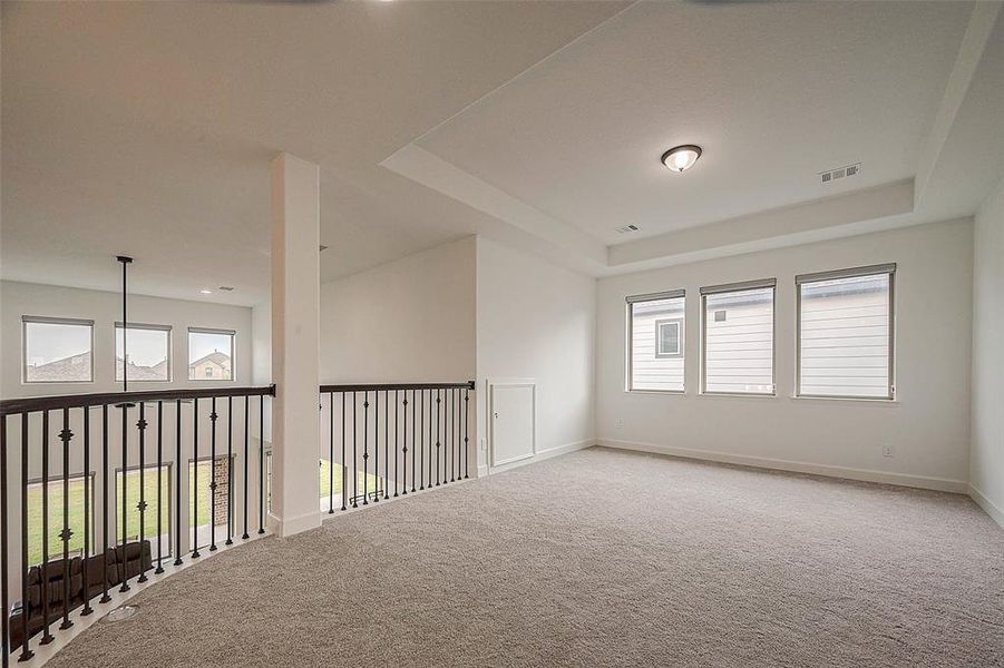 This is a spacious, carpeted room with natural light from three windows. It features a modern wrought-iron railing overlooking the lower level, and a neutral color palette perfect for personalizing.