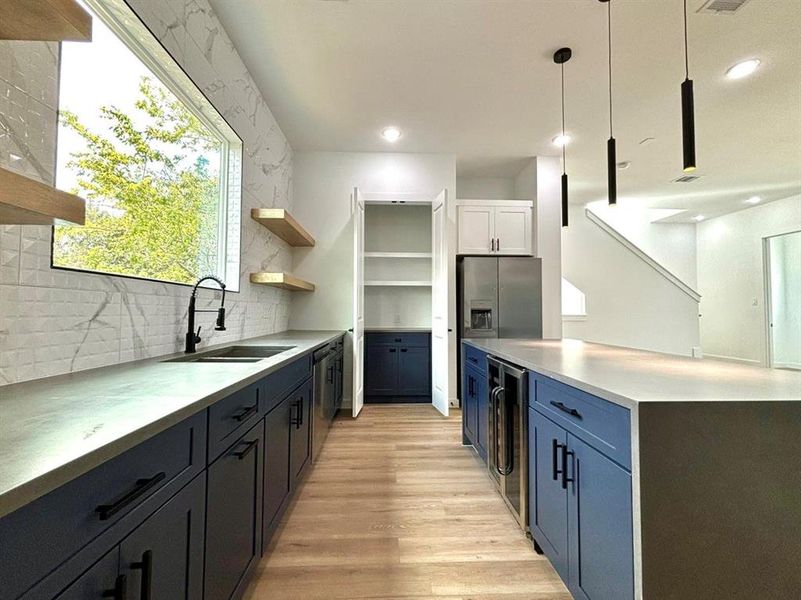 This is a bright and modern kitchen featuring sleek dark blue cabinetry, contrasting gray countertops, and stylish floating shelves. Natural light streams and elegant pendant lights complement the elegance of this kitchen. The room is finished with luxury vinyl flooring, giving it a warm, inviting feel.