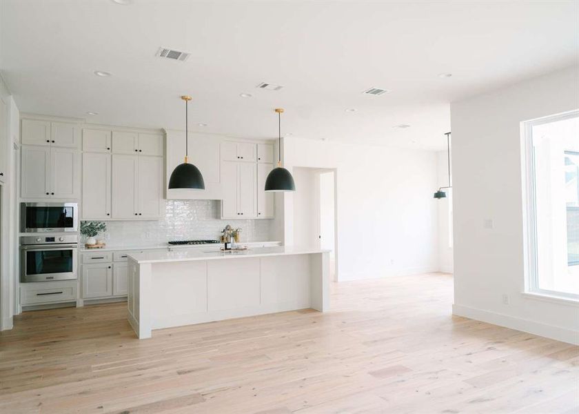 Kitchen with light hardwood / wood-style flooring, appliances with stainless steel finishes, an island with sink, decorative light fixtures, and tasteful backsplash