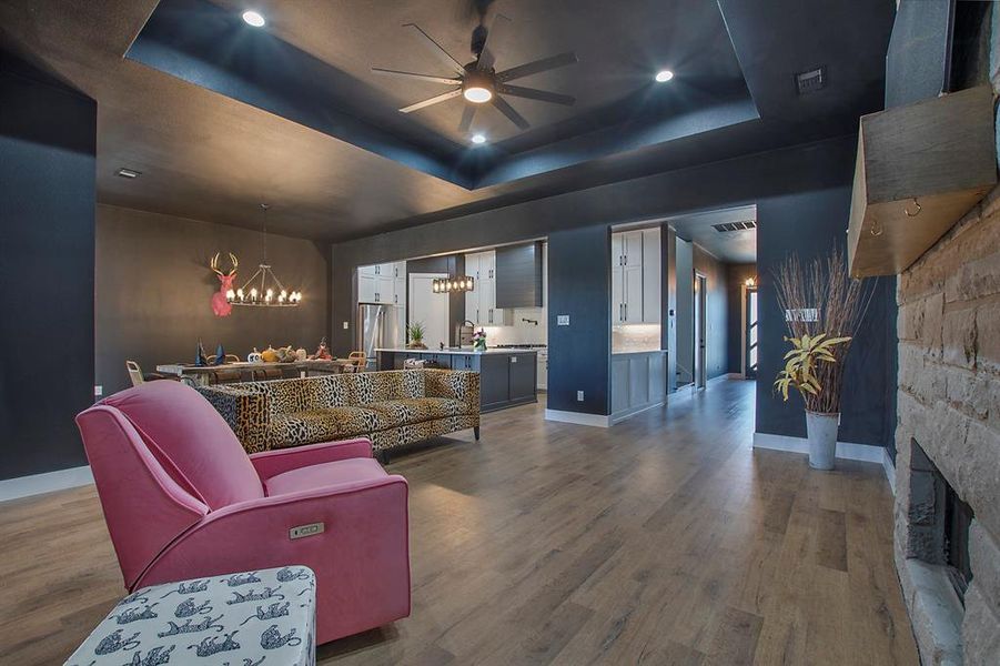 Living room with a stone fireplace, hardwood / wood-style floors, ceiling fan with notable chandelier, and a raised ceiling