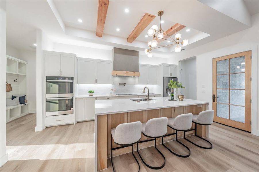 Kitchen featuring wall chimney range hood, stainless steel appliances, a kitchen island with sink, beam ceiling, and blonde plank, wood-like luxury vinyl floors.