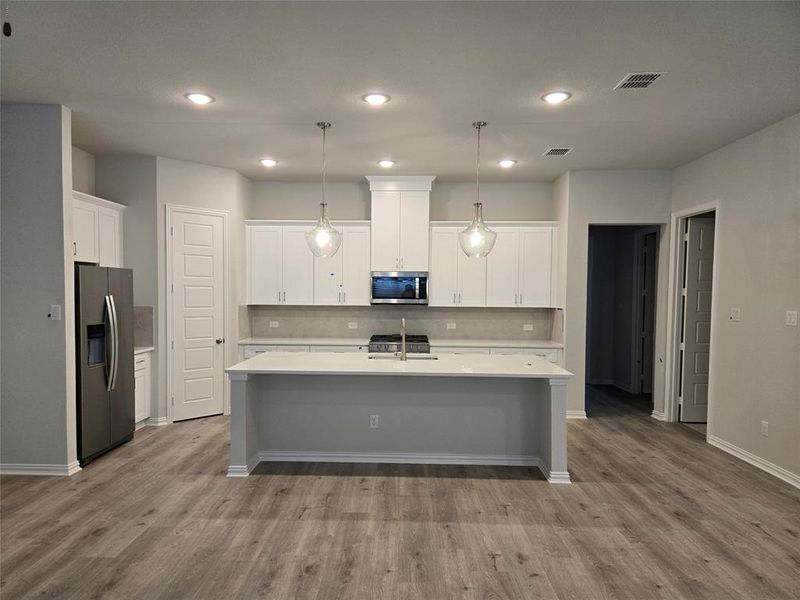 Kitchen with light hardwood / wood-style floors, hanging light fixtures, white cabinets, appliances with stainless steel finishes, and an island with sink